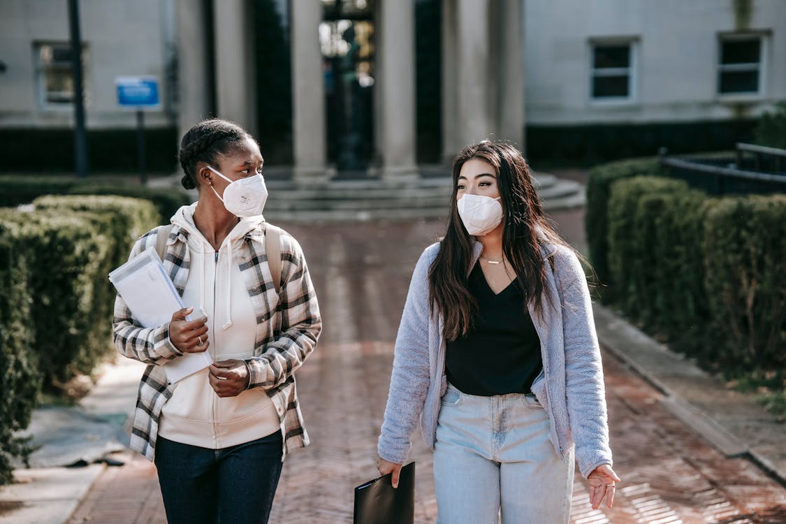 Diverse women in respirators with folders walking on campus sidewalk