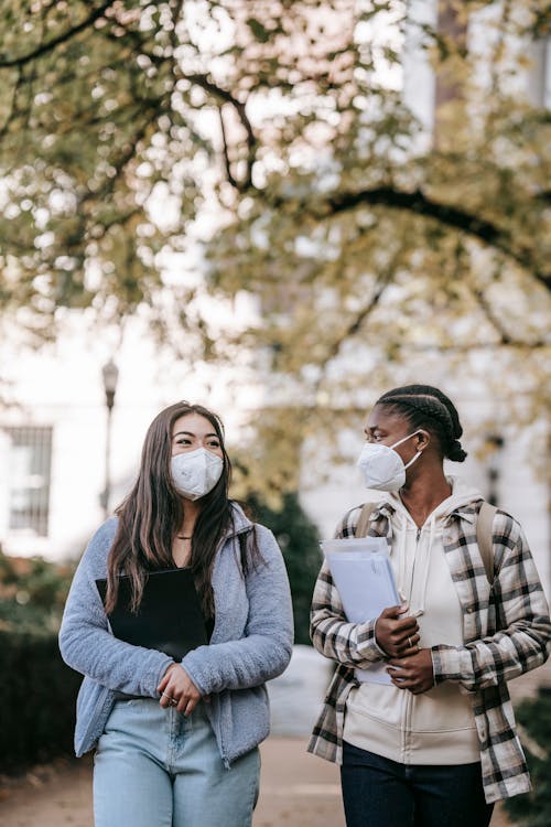 Content young multiracial female students in casual outfits and protective masks carrying folders and having pleasant conversation while walking together on city street