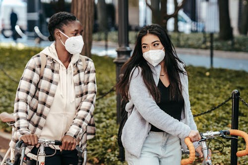 Free Multiethnic girlfriends in masks talking to each other while strolling with bicycles in park during COVID pandemic Stock Photo