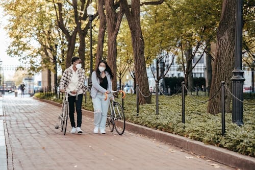 Full body of diverse women in masks with bicycles walking on paved walkway along trees in park during coronavirus period