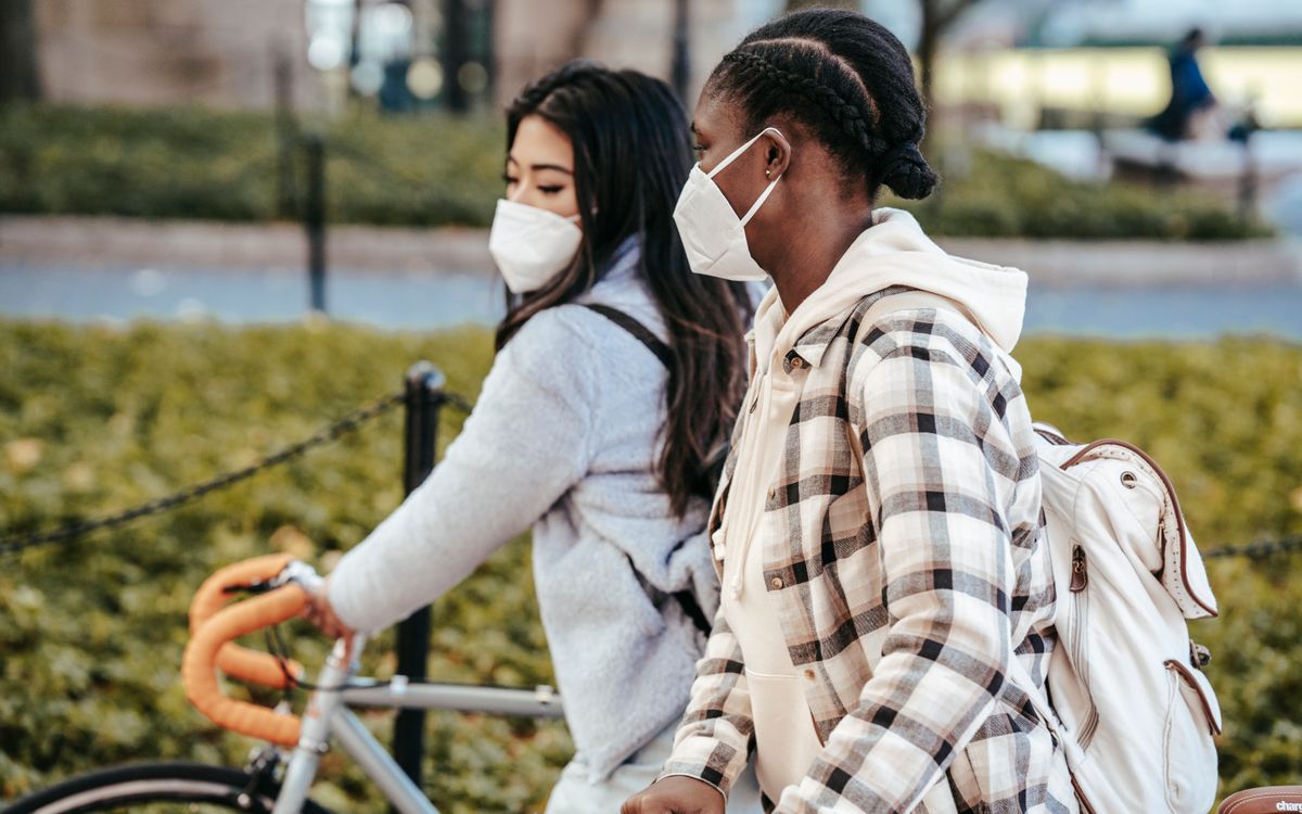 Side view of multiethnic female friends in casual clothes and masks protecting from coronavirus walking with bicycles in park