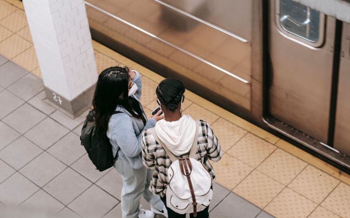 From above back view of unrecognizable black female student with girlfriend in mask in subway against public transport