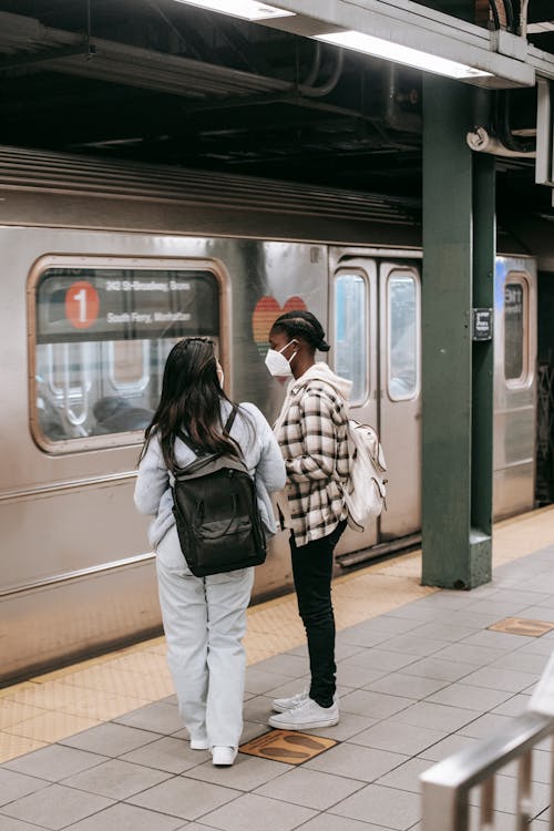 Back view of anonymous multiracial girlfriends in casual clothes with backpacks in masks against public transport in subway station
