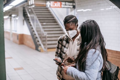 Free Unrecognizable young multiethnic girlfriends in masks touching screen on cellphones while speaking on platform of underground Stock Photo