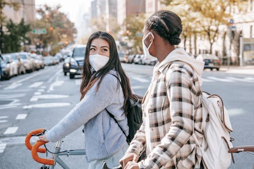 Side view of unrecognizable multiethnic girlfriends in respiratory masks with bicycles talking and looking at each other on urban roadway