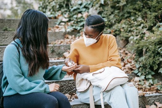 Unrecognizable ethnic student applying sanitizer on hand of girlfriend outdoors