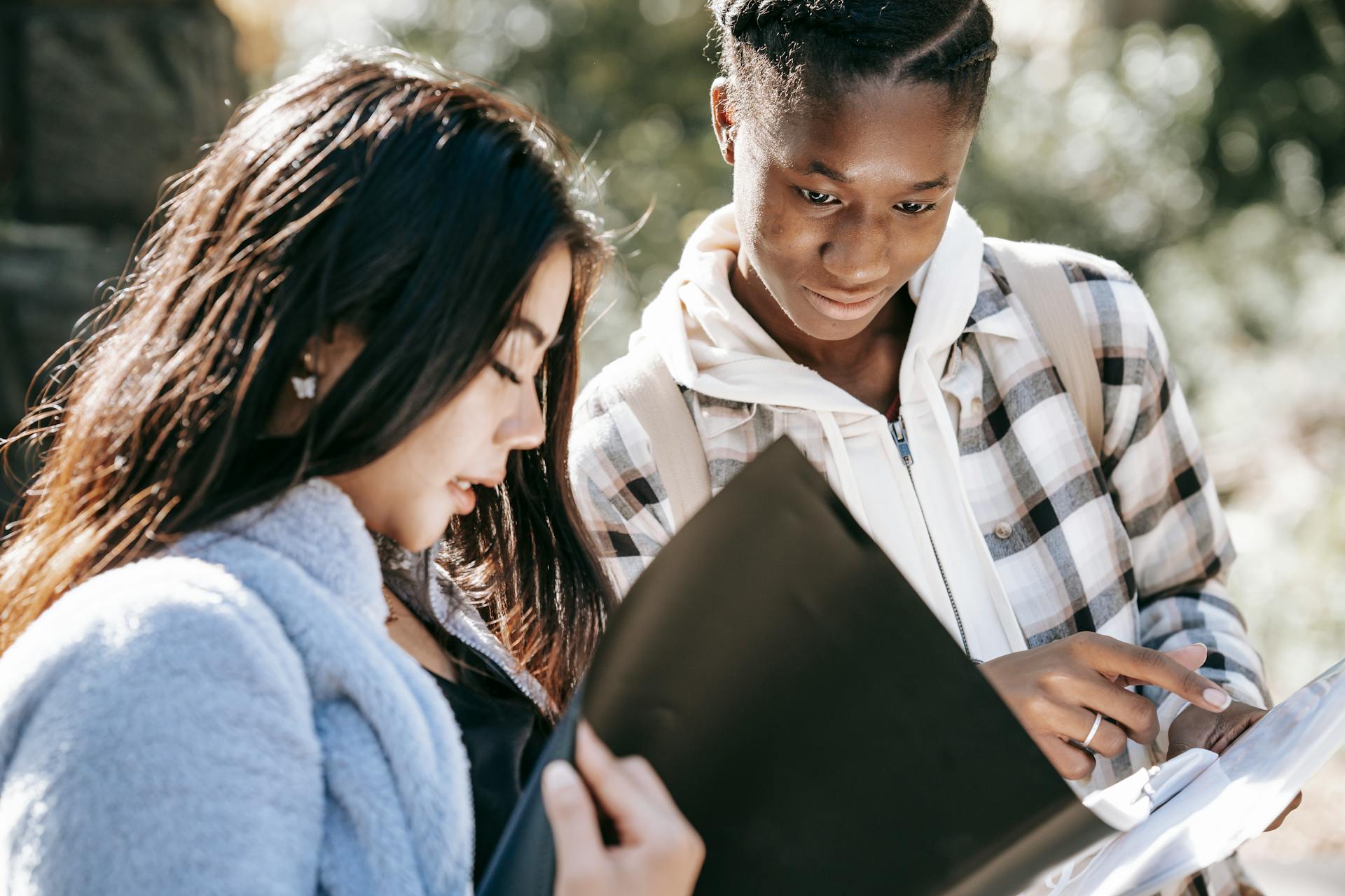 Crop young multiracial best female friends sharing folders while preparing homework and talking in back lit