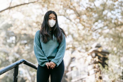 一位年轻、沉思的少数族裔女性身穿休闲服装，头戴呼吸面罩，双手紧握，低头俯视着城市