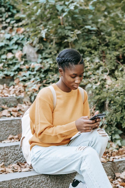 Young focused African American female with backpack surfing internet on mobile phone while resting on city staircase in fall