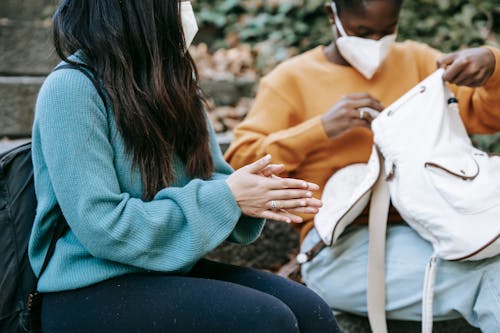 Crop anonymous multiracial best female friends in masks with backpacks speaking on urban stairs during coronavirus pandemic