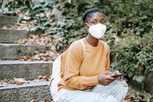 Anonymous young African American female in respiratory mask and casual wear with cellphone looking forward while sitting on city stairs