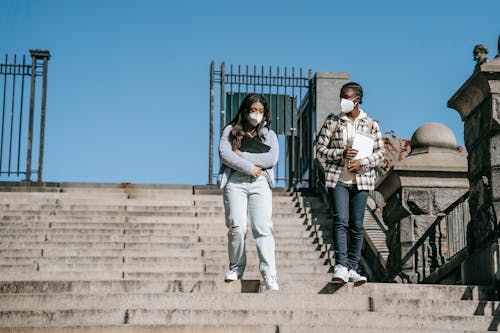 Unrecognizable young multiethnic female students in respiratory masks talking while strolling on city staircase under blue sky