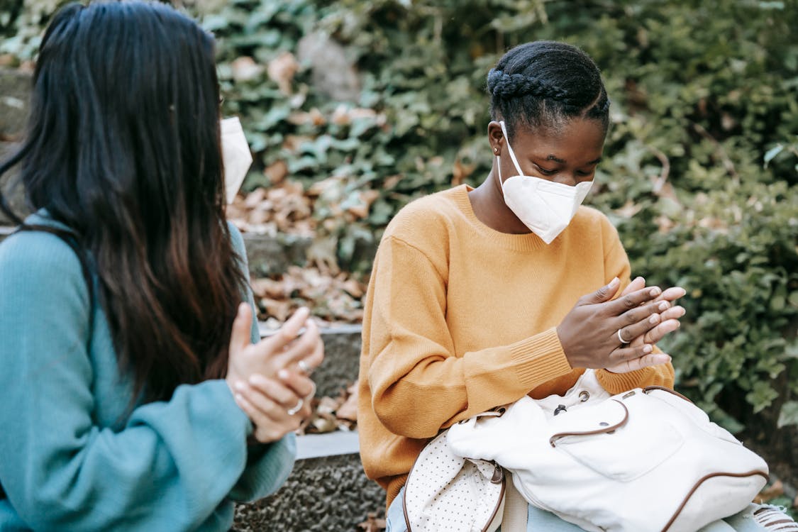 Young multiracial female friends in casual clothes and protective face masks wiping hands and sitting on stone stair in park