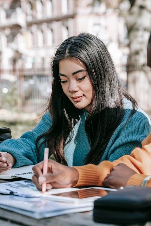 Content young multiracial female students in warm sweaters working on university project together and taking notes in copybooks while sitting at table in park