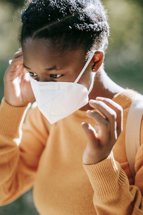 Crop calm young African American female in yellow sweater putting on face mask and looking away while standing on blurred park background on sunny autumn day