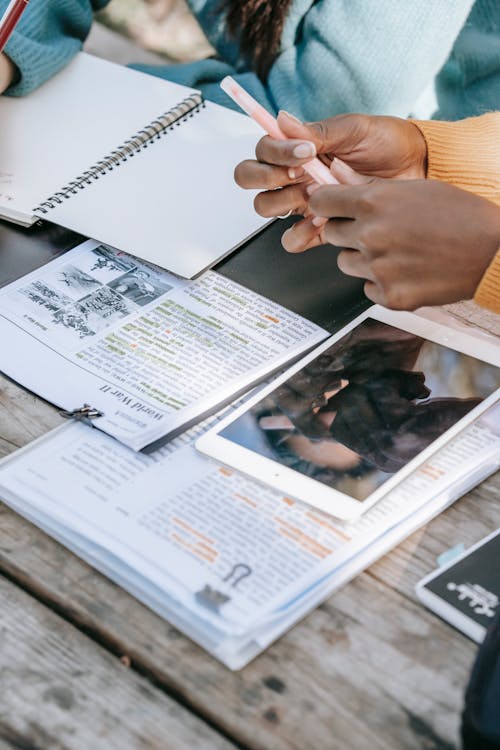 Free From above crop anonymous multiracial female students sitting at table with textbook copybook and tablet while working on home assignment together in sunny park Stock Photo