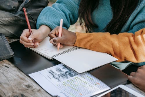 Crop unrecognizable diverse schoolgirls taking notes in copybook in park