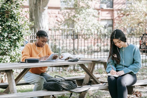 Positive young multiracial female students sitting at table in city park and preparing for exams together while browsing tablet and taking notes in copybook