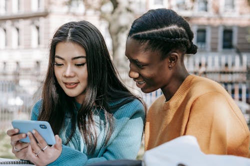 Young diverse cheerful friends smiling and browsing mobile phone on blurred background of street