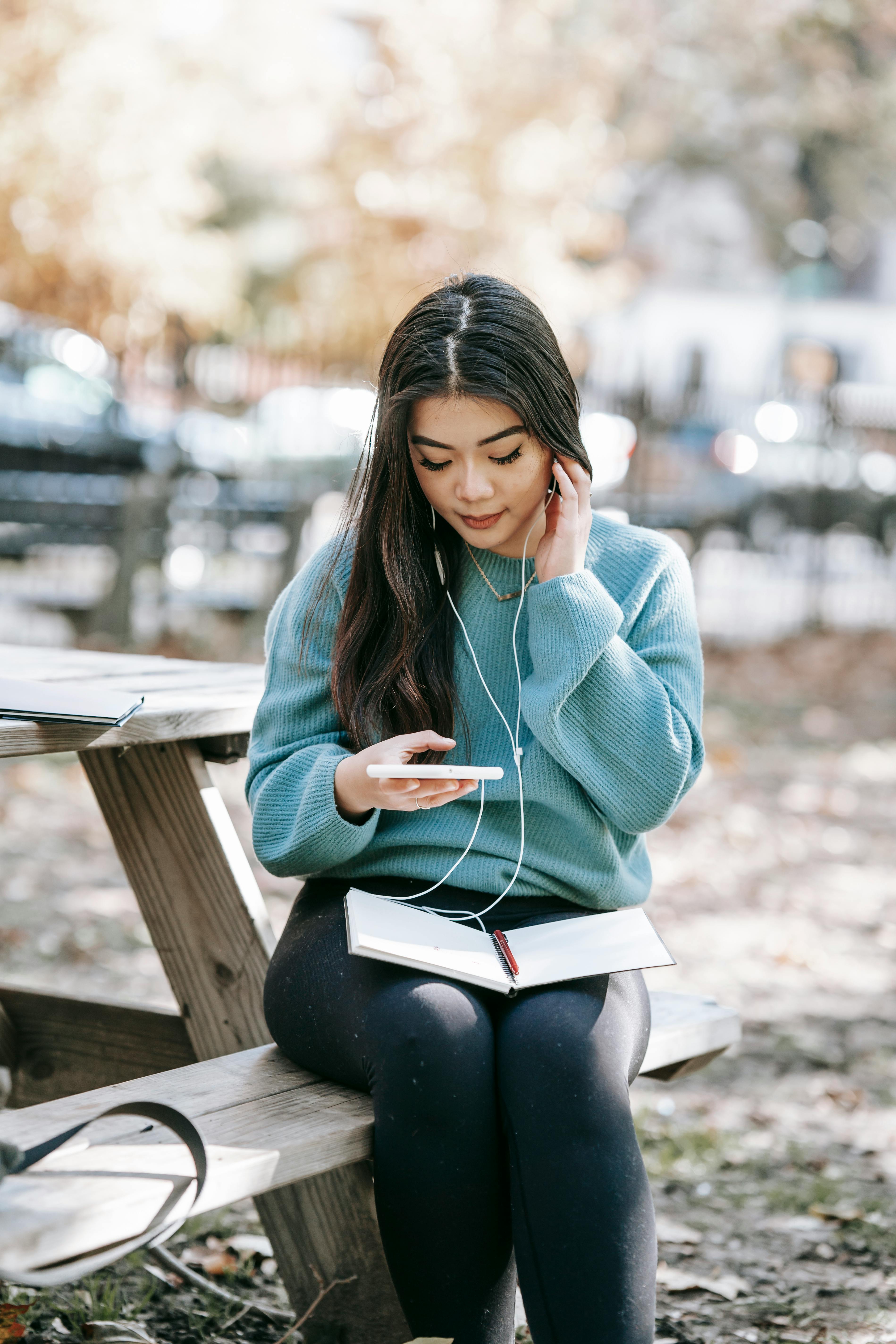 young asian woman in earphones with notebook using smartphone