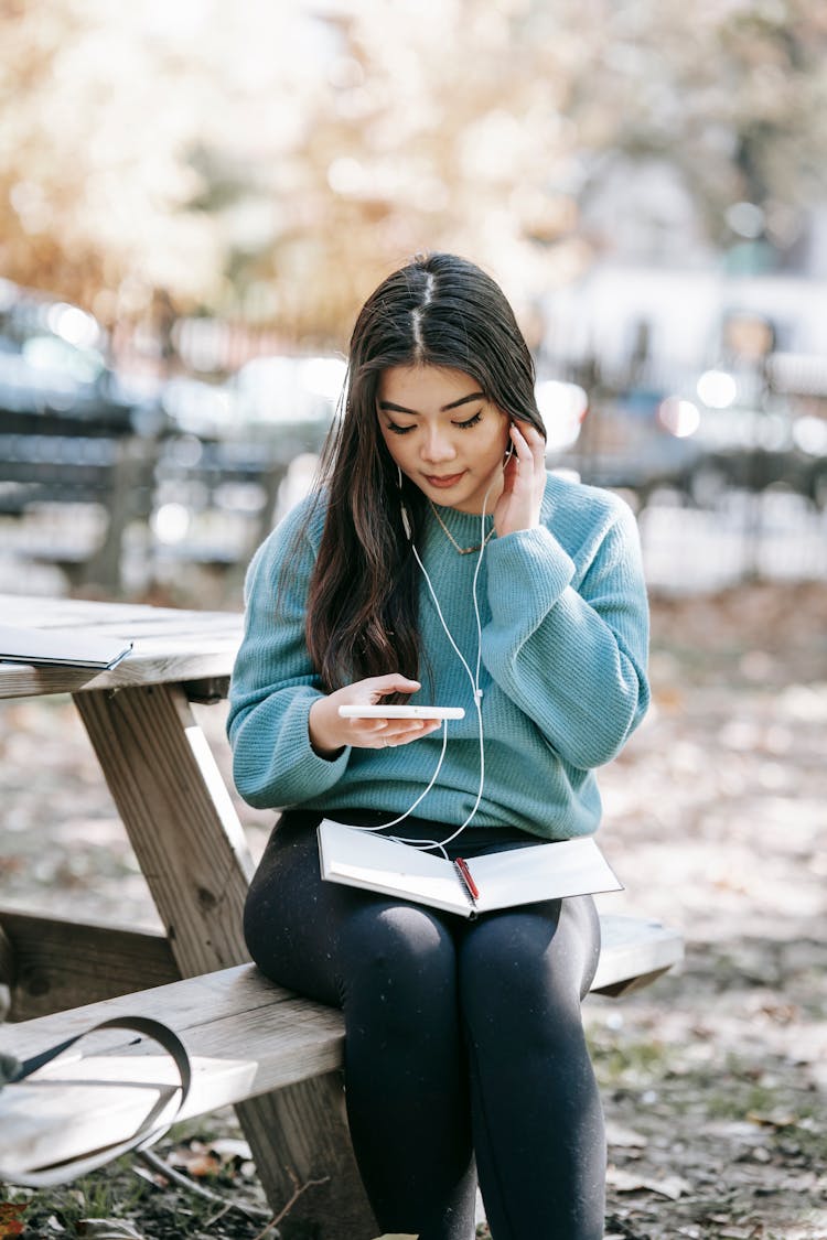 Young Asian Woman In Earphones With Notebook Using Smartphone