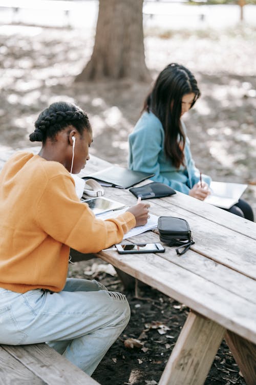 Focused anonymous students in casual outfit writing report in notebooks at table in park