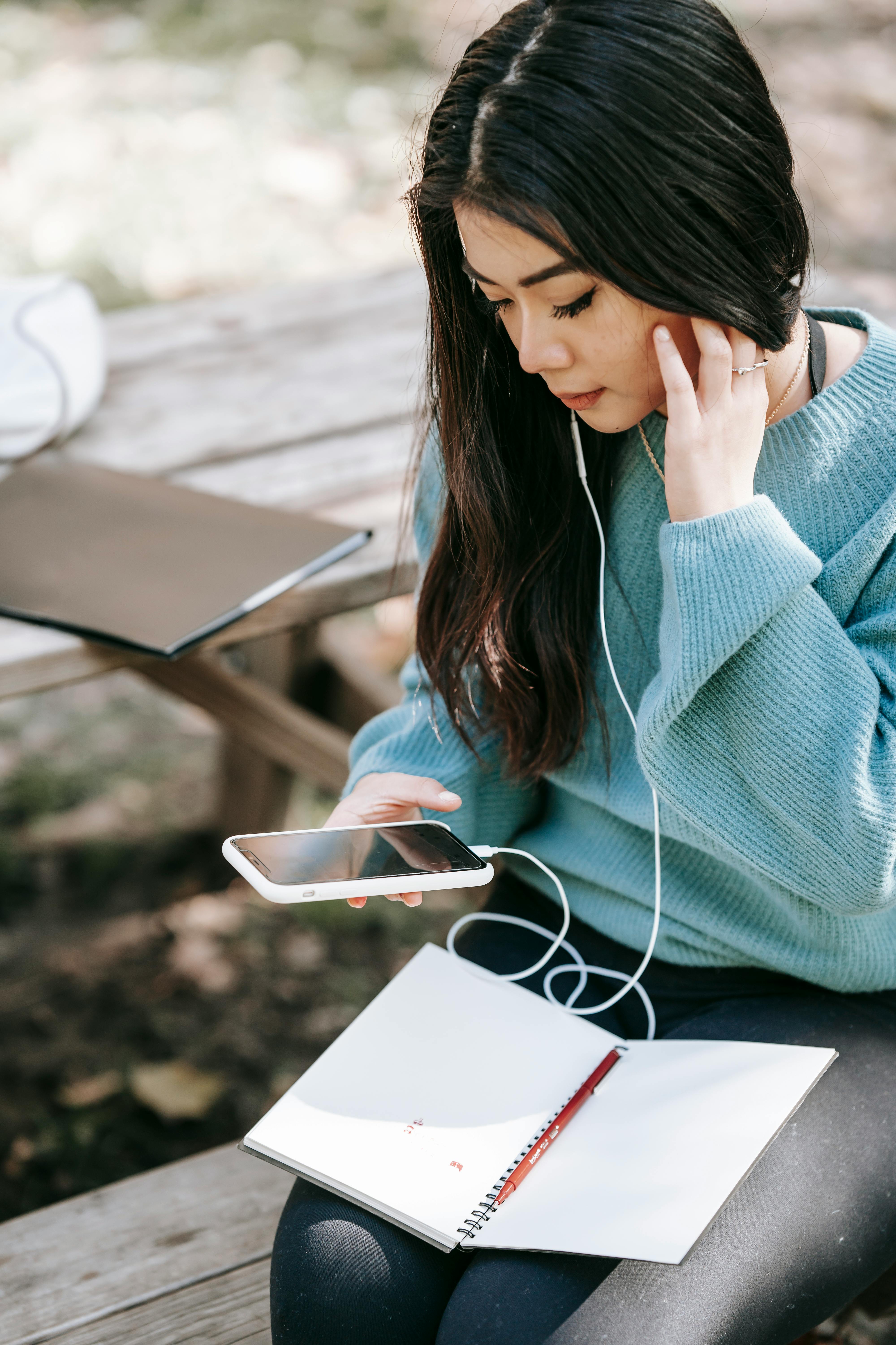 young asian woman in earphones with notebook using smartphone