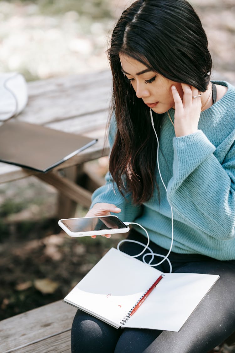 Young Asian Woman In Earphones With Notebook Using Smartphone