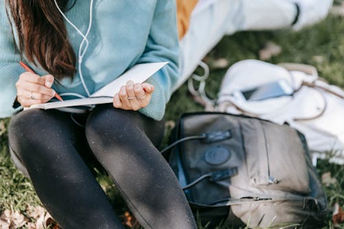 Crop anonymous female listening to music in earphones while writing in notebook near bags on grass