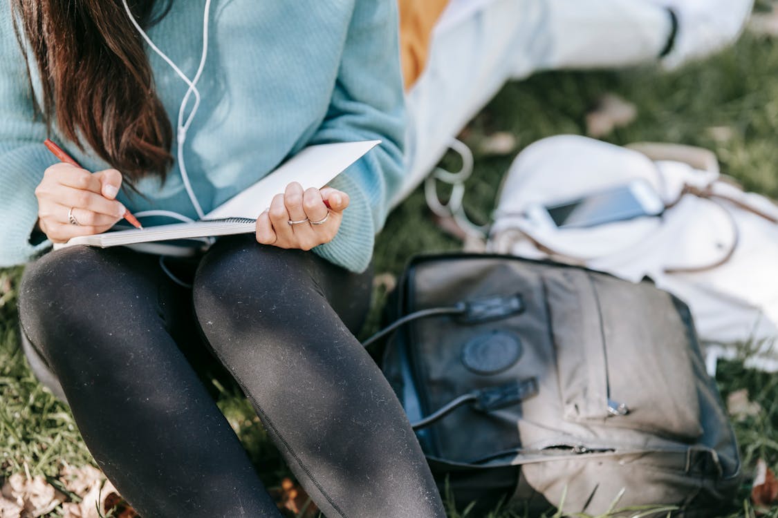 Free Crop anonymous female listening to music in earphones while writing in notebook near bags on grass Stock Photo