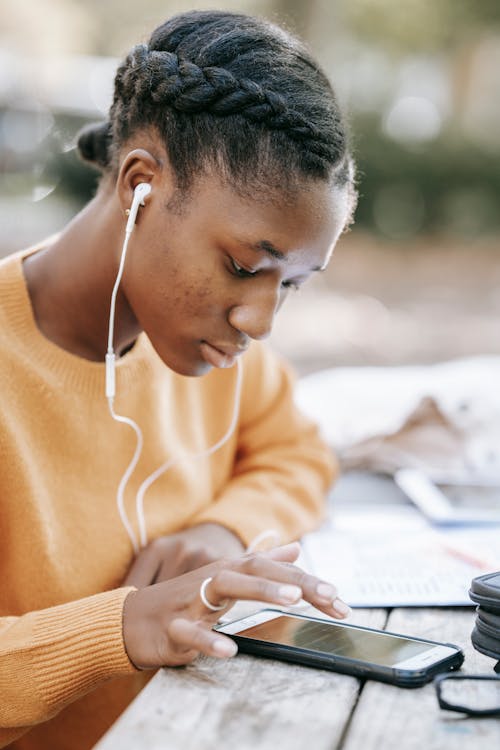 Young African American female using and checking smartphone while listening music with earphones at table