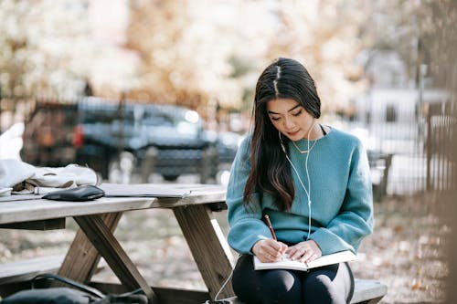 Young focused Asian woman in earphones writing in diary