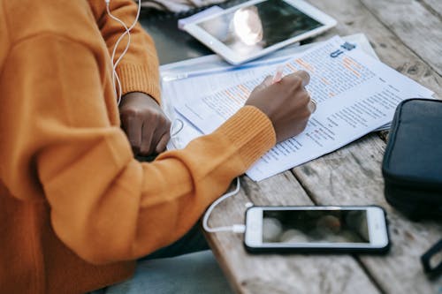 From above of crop anonymous African American female in sweater studying and outlining words on paper at table with smartphone