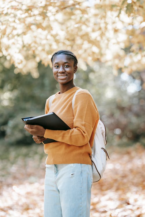 Free Young black happy woman with folder and backpack near tree Stock Photo