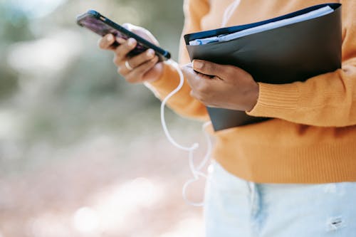 Crop anonymous African American female in warm sweater with folder touching screen of mobile phone on blurred background