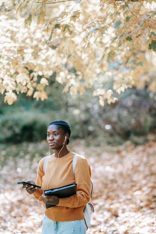 Young African American female with folder looking away and using earphones with mobile phone while listening to music near tree