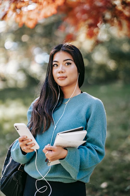 Content young ethnic female in sweater listening podcast with earphones and mobile phone on blurred background of park