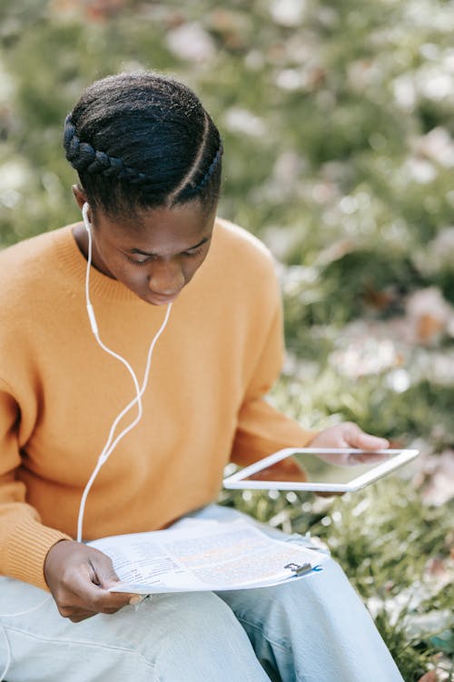 High angle of young focused African American female student in earphones doing homework using tablet in garden