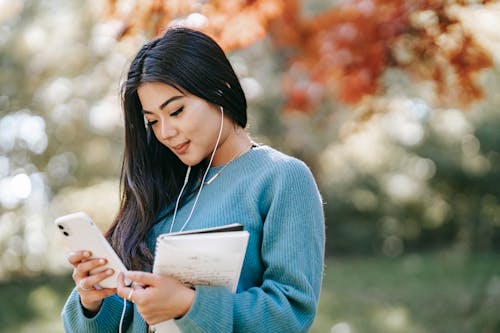 Side view of young smiling Asian female student with notebook and earphones browsing mobile phone in park