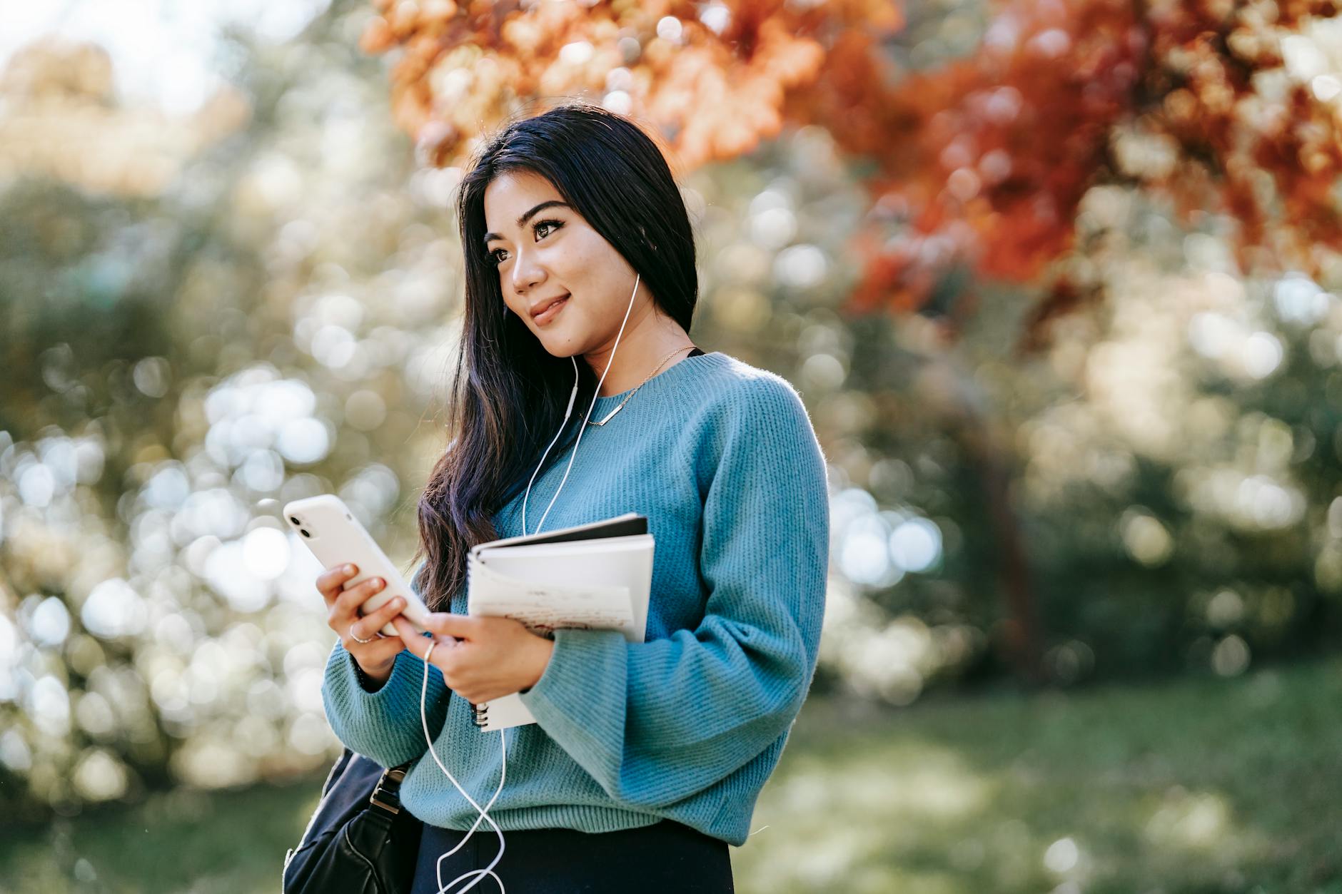 Cheerful young woman outdoors, using phone and earbuds, holding notebook, relaxing in a park.