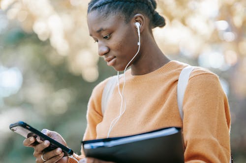 Low angle of young African American female student in casual clothes and earphones browsing mobile phone on street