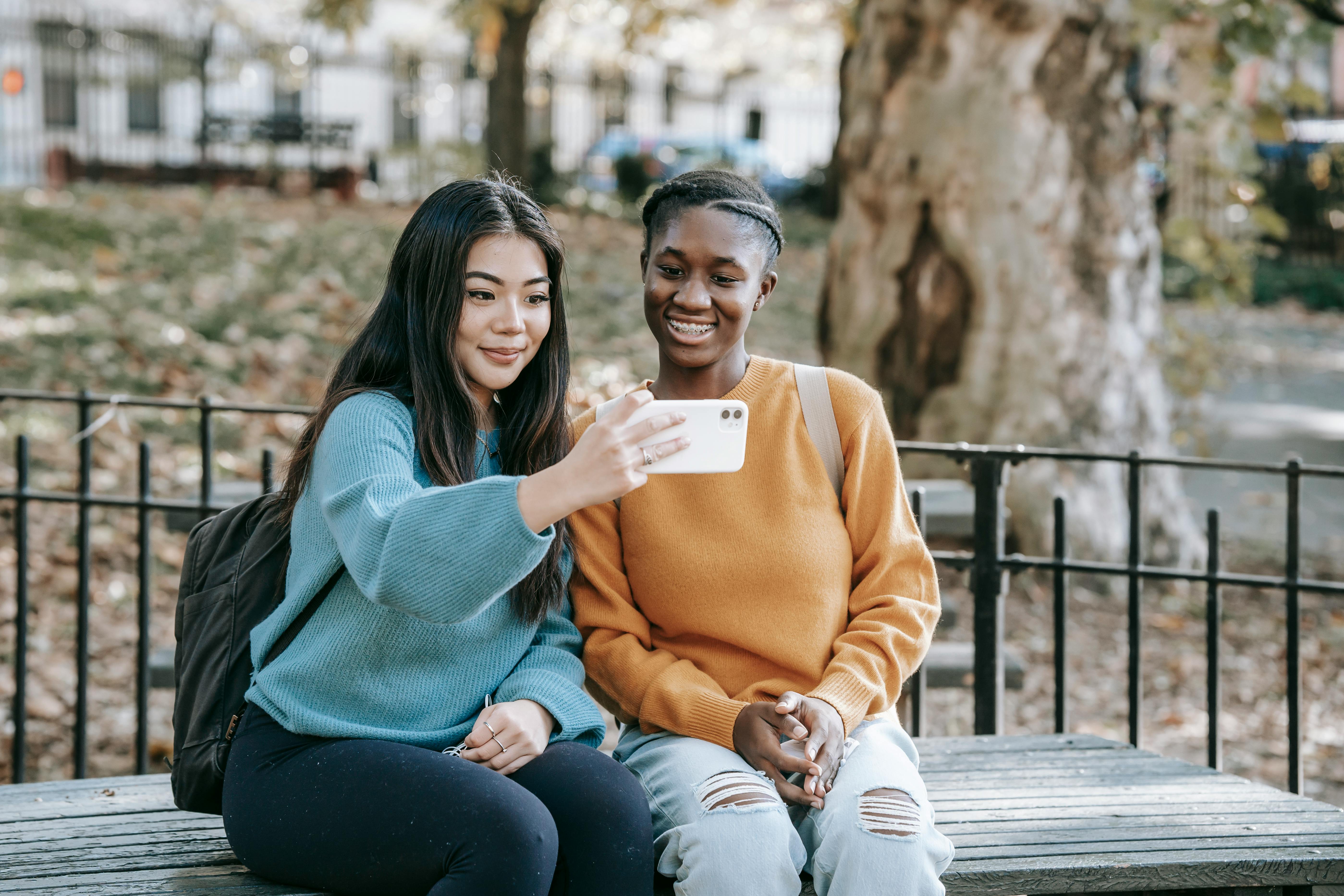 happy young friends using smartphone sitting on bench in garden