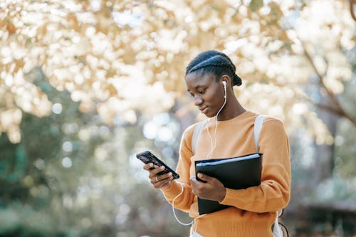 Positive young African American female student with earphones and folder using mobile phone in park