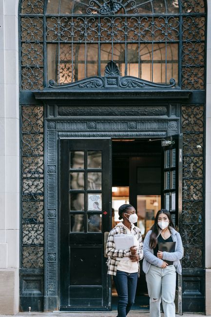 Young multiethnic students in protective masks with papers standing against entrance of university with massive door