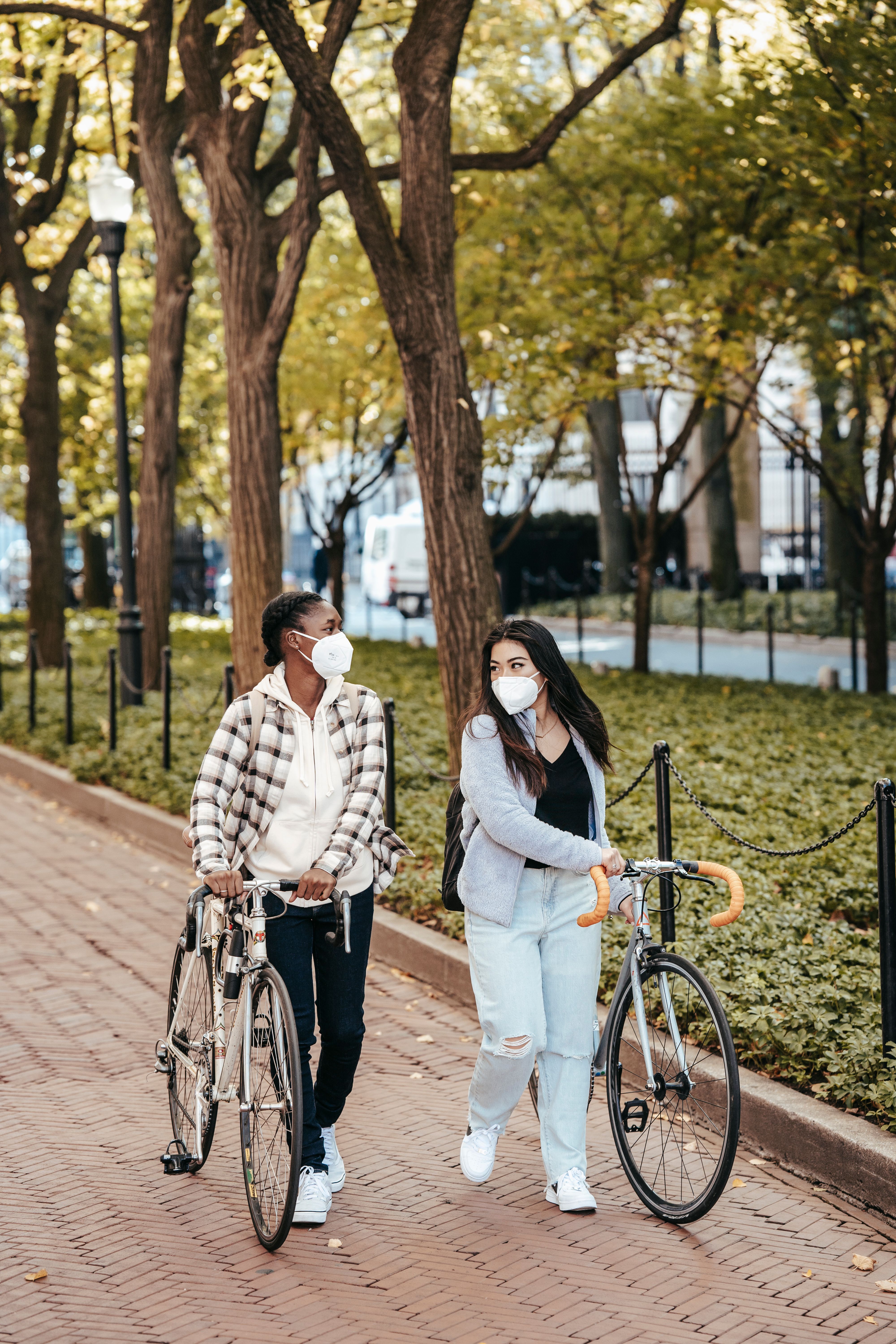 白天，两名戴着口罩的妇女在绿树成荫的公园里骑着自行车散步