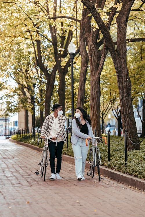 Full body of multiracial girlfriends in trendy clothes and masks strolling on pavement with bikes and looking at each other