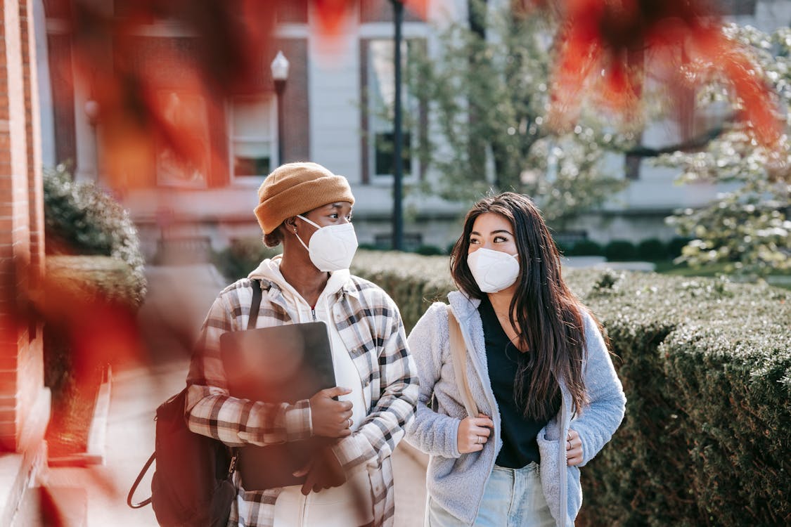 Young multiracial girlfriends in casual clothes with masks on faces strolling together in autumn park