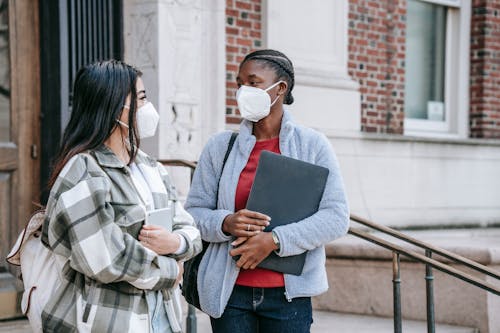 Young multiracial women in masks with laptop and tablet standing on stair and communicating while looking at each other