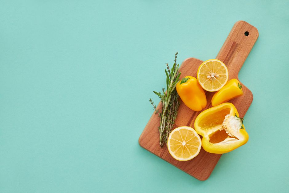 A vibrant display of yellow peppers, lemons, and herbs on a wooden board against a teal background.