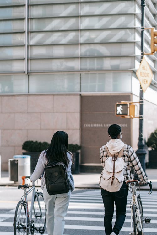 Back view of unrecognizable girlfriends with backpacks crossing asphalt road with bikes while spending time together